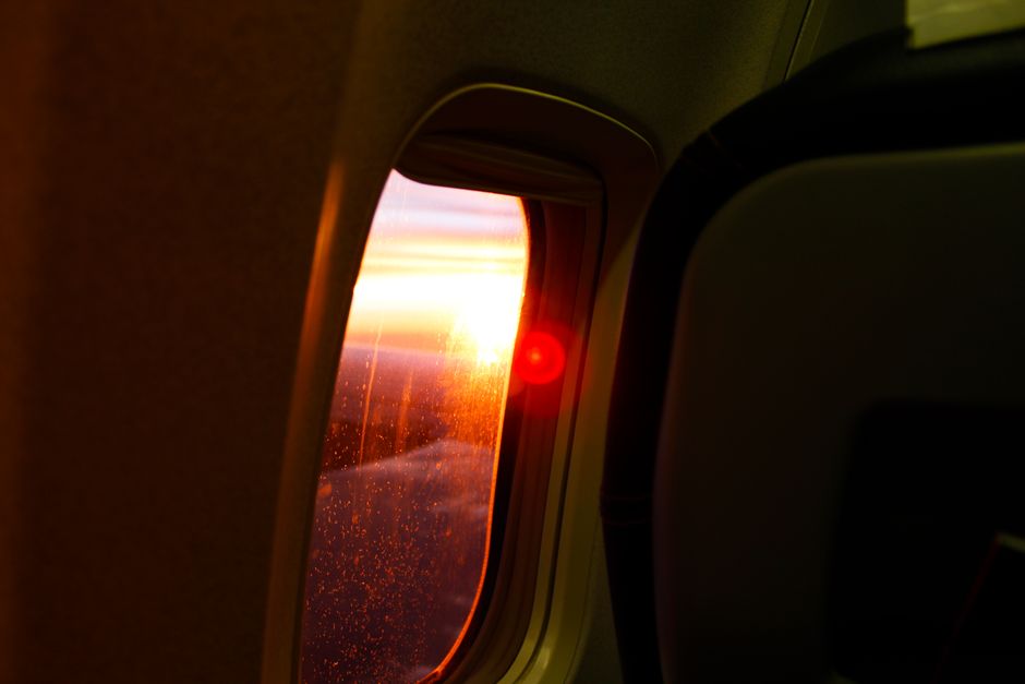 A passenger looking out an airplane window at the sunrise over clouds after an overnight red-eye flight, hinting at arrival.