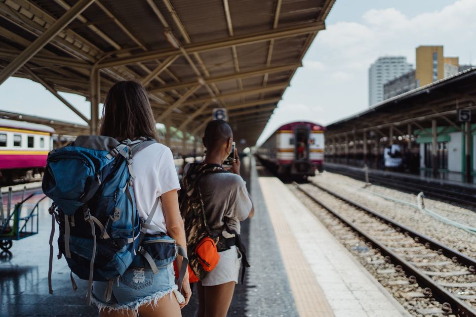 Two backpackers looking at a large train schedule board inside a historic European train station, with their backpacks at their feet.