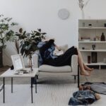 A person in professional attire looking overwhelmed by a cluttered living room with clothes on chairs and papers on a coffee table, signifying the dread of cleaning day.