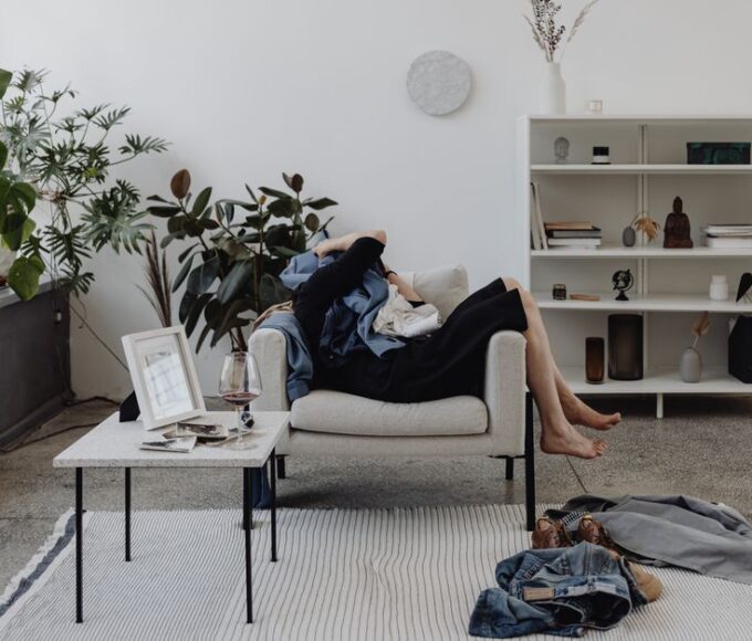 A person in professional attire looking overwhelmed by a cluttered living room with clothes on chairs and papers on a coffee table, signifying the dread of cleaning day.