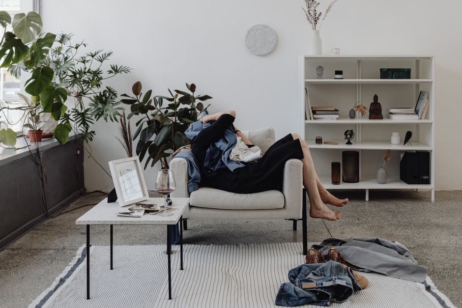 A person in professional attire looking overwhelmed by a cluttered living room with clothes on chairs and papers on a coffee table, signifying the dread of cleaning day.