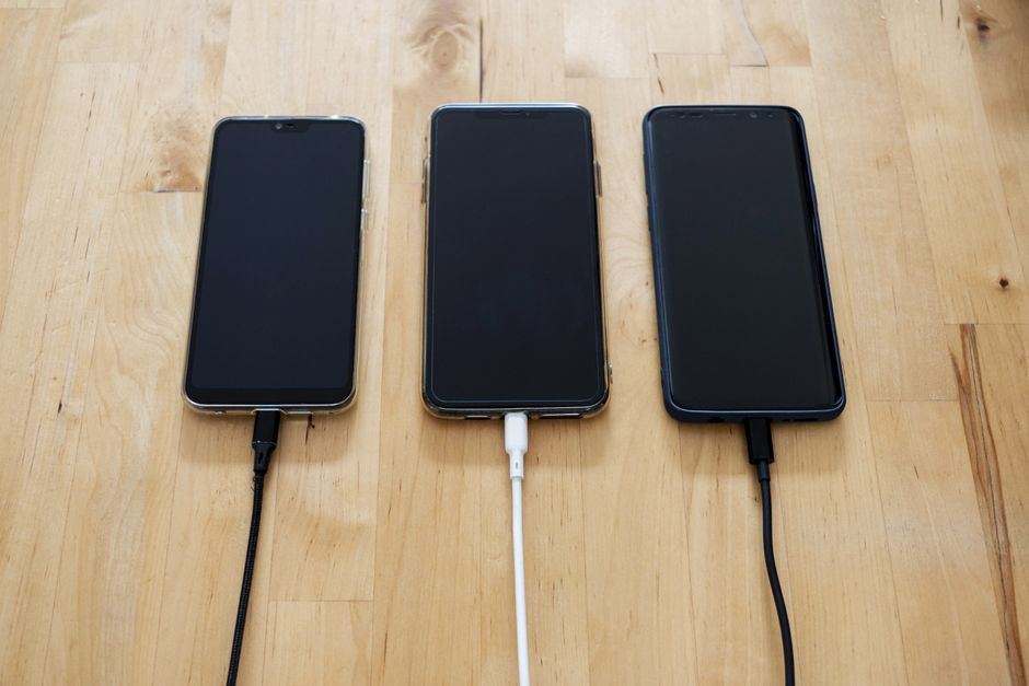 A hand connecting a USB-C cable from a large power bank to a smartphone, with other travel gadgets like earbuds and a tablet in the background, showing multi-device charging during a trip.
