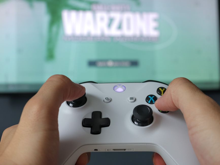 A close-up shot of a modern console controller resting on a table, with the vibrant, out-of-focus menu of a free-to-play game visible on a screen in the background, subtly highlighting in-game currency without aggressive monetization.
