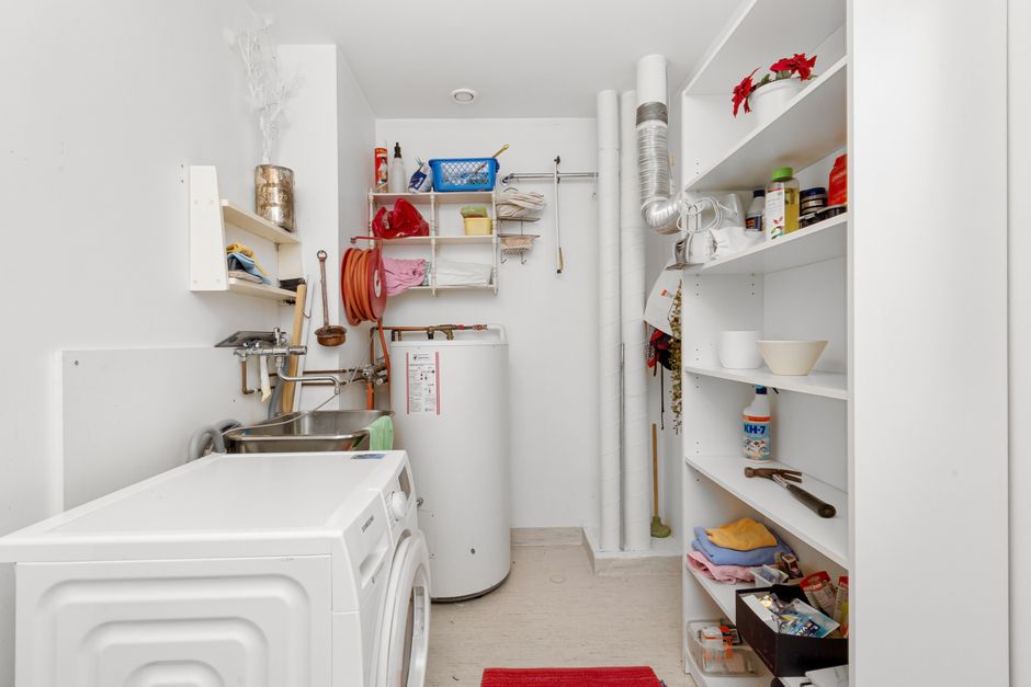 A collapsible, multi-tier drying rack fully extended in a corner of a small apartment, holding various items of clothing, with a window nearby for ventilation.