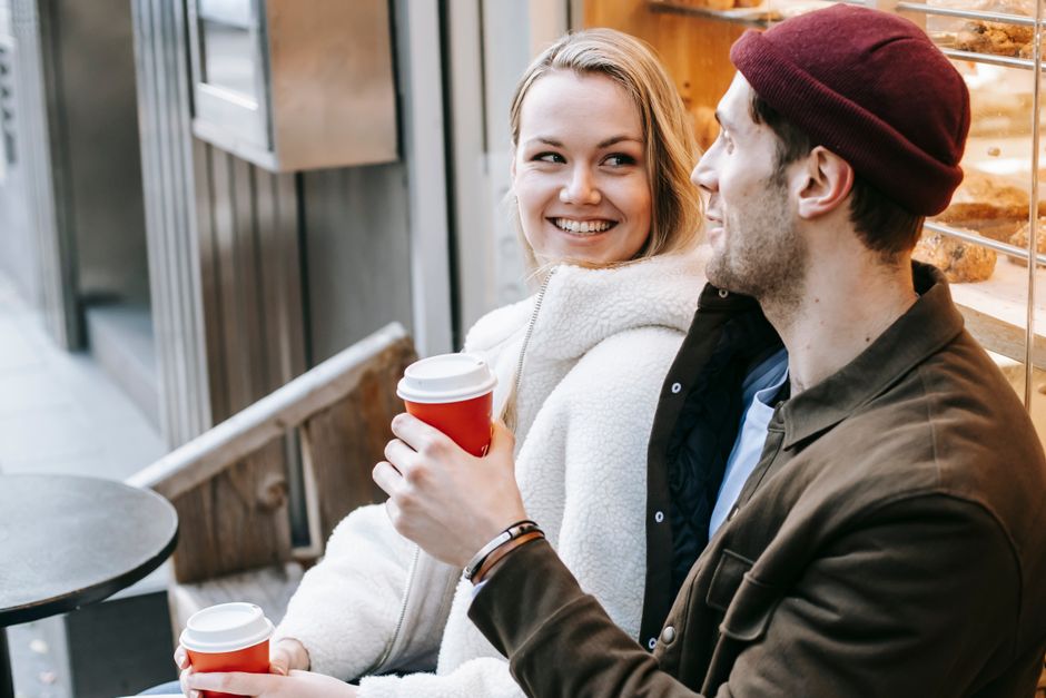 Two young professionals having coffee outdoors, dressed in layered, versatile outfits suitable for varying temperatures and urban environments.