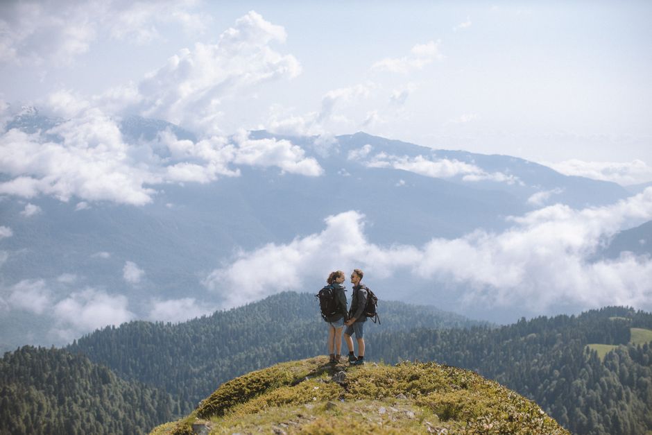 A couple in hiking gear on a mountain trail, overlooking a vast, scenic landscape with blue skies and distant peaks, illustrating active exploration.