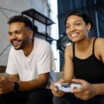 A couple on a couch, one holding a console controller, the other watching intently, both smiling.