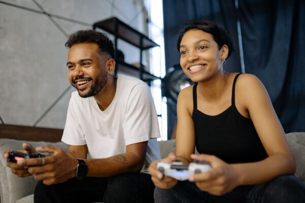 A couple on a couch, one holding a console controller, the other watching intently, both smiling.