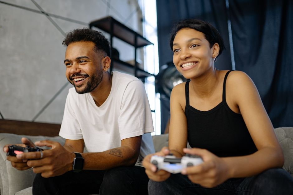 A couple on a couch, one holding a console controller, the other watching intently, both smiling.