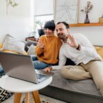 A couple on separate laptops, one in a city apartment, the other in a cozy home, having a video call with a split screen, symbolizing long-distance communication.