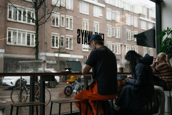 A person working on a laptop in a warmly lit cafe, looking out at a rainy city street.