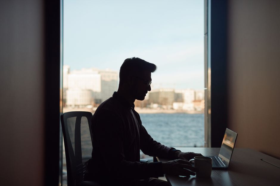 A person working on a laptop on a balcony with a panoramic view of Lisbon's red-tiled roofs and the Tagus River.
