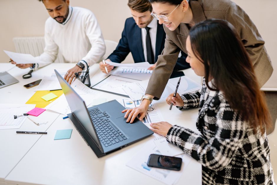 Three professionals from diverse backgrounds discussing documents and working on a laptop, illustrating global collaboration on AI regulation