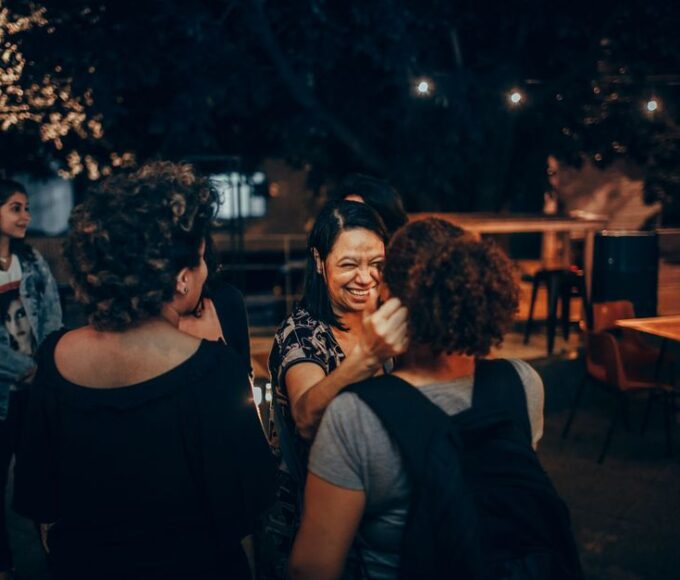 A diverse group of young adults, possibly expats, laughing and chatting around a table at an outdoor café in a sunny European city.