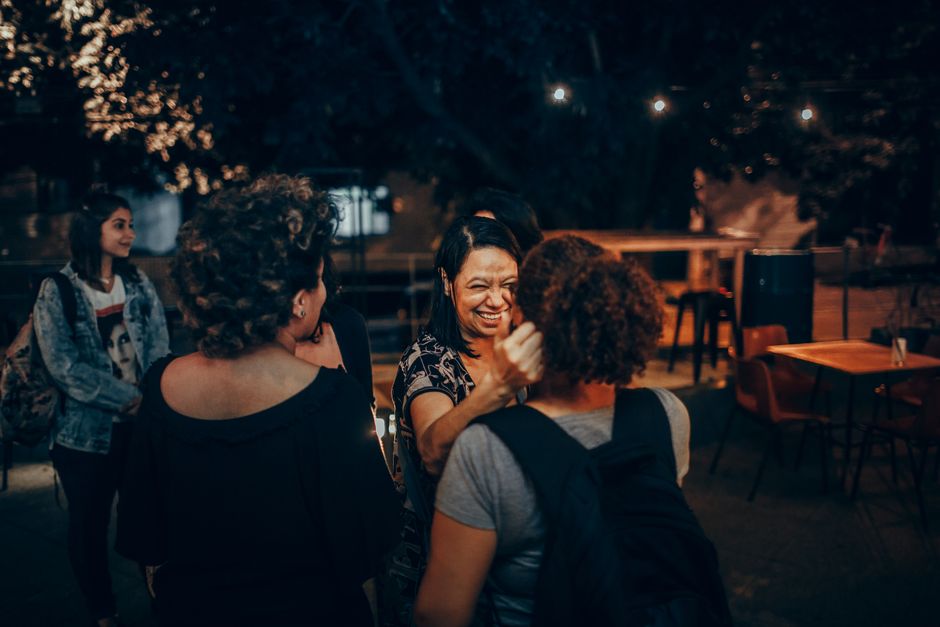 A diverse group of young adults, possibly expats, laughing and chatting around a table at an outdoor café in a sunny European city.