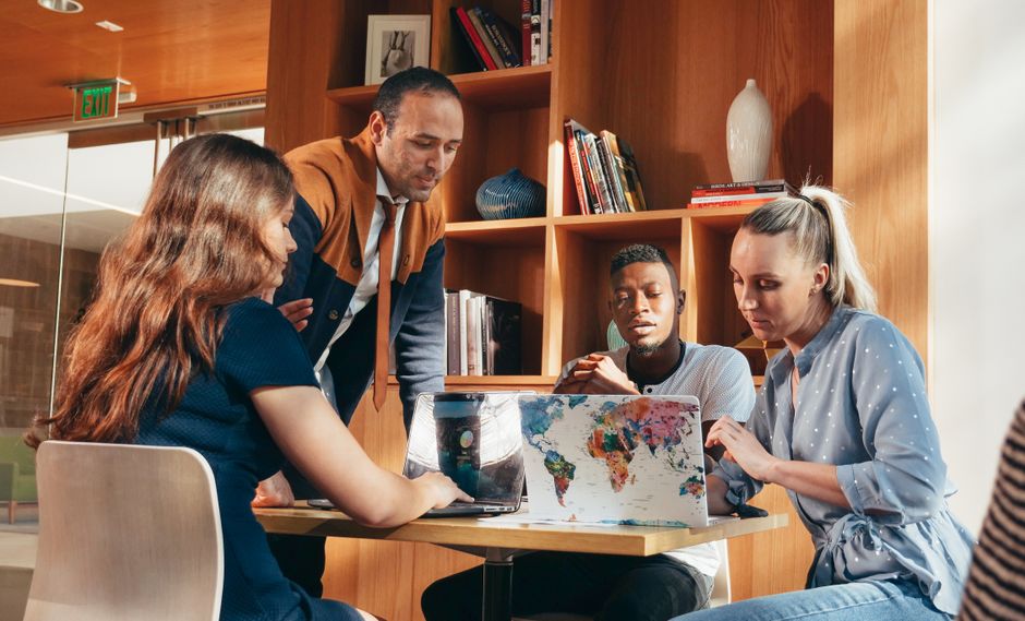 Three people, diverse in age and ethnicity, are gathered around a table with laptops and papers, engaged in a discussion, symbolizing collaborative information seeking.