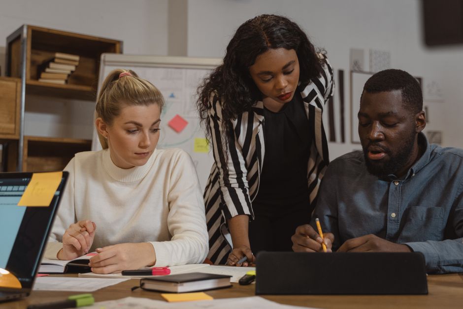 A group of people of varying ages and backgrounds engaged in conversation around a tablet, with one person gesturing to explain a concept, representing the challenge of explaining AI to a non-technical audience.