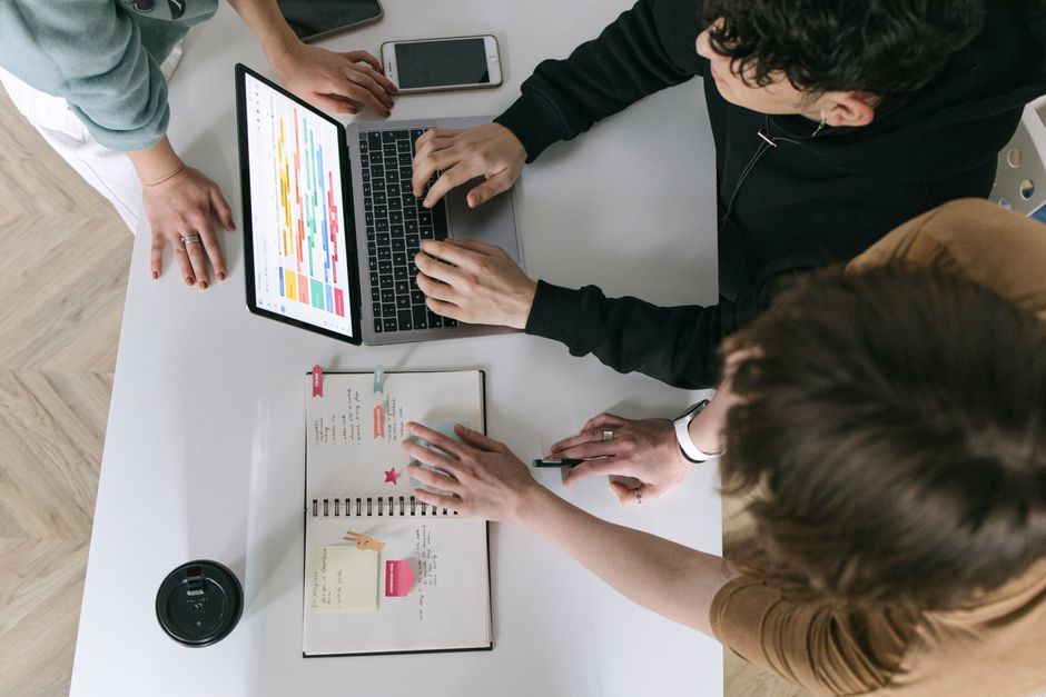 Three diverse individuals from different continents (representing North America, Europe, South America) collaboratively viewing a large digital calendar on a screen, making adjustments.