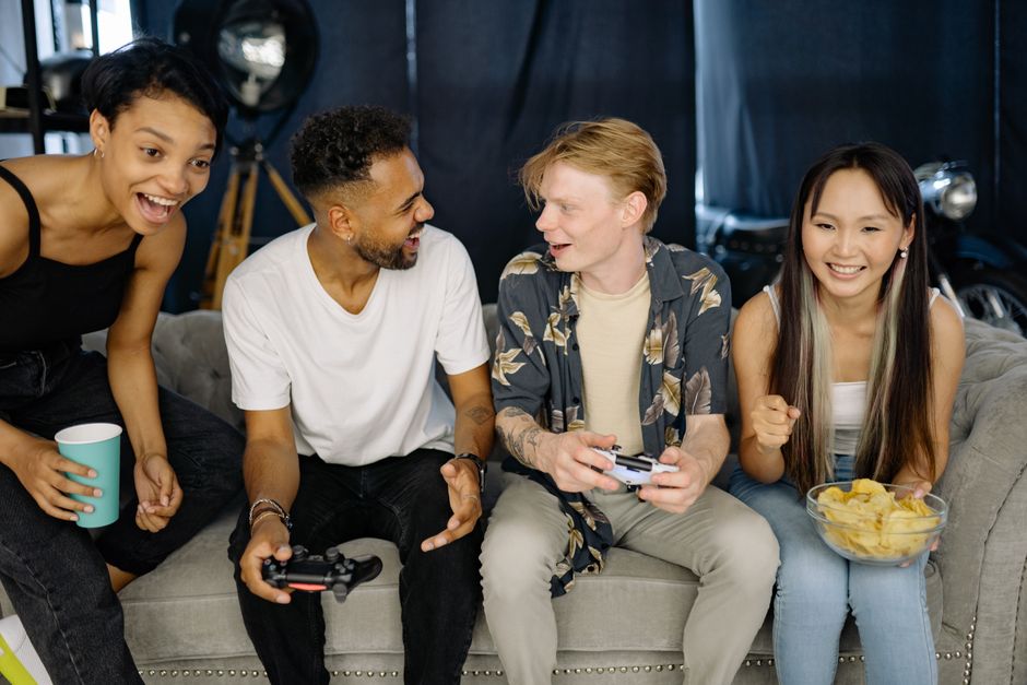 A diverse group of adults and teenagers smiling while playing video games together on different screens.