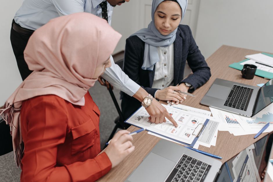 A small business team, ethnically diverse, gathered around a tablet, discussing AI-generated reports and reviewing data with human oversight.