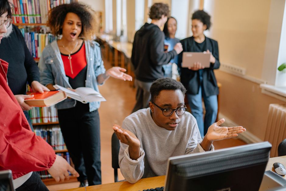 A group of diverse students from Europe and the Americas actively collaborating around a table in a university common area, with various digital devices and notes, representing teamwork and tech use.
