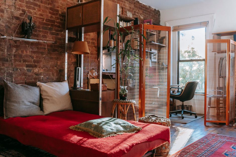 An eclectic bedroom with a mix of secondhand furniture, including a wooden dresser and a metal bed frame, adorned with patterned textiles and various green plants.