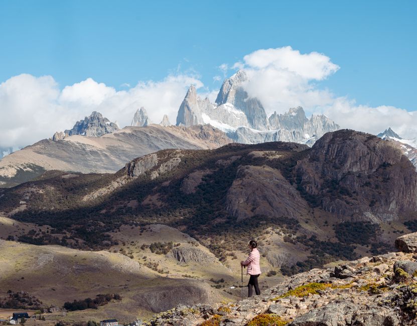 Two hikers, a man and a woman, stand on a rocky trail with the majestic granite spires of Fitz Roy and its surrounding peaks in El Chaltén, Patagonia, visible in the background under a dramatic sky.