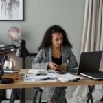 An expat woman with a laptop, budgeting, with a global map and currency symbols in the background.