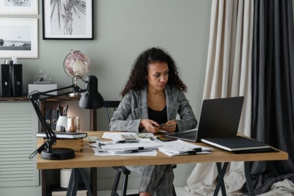 An expat woman with a laptop, budgeting, with a global map and currency symbols in the background.