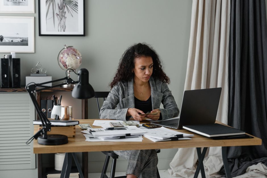 An expat woman with a laptop, budgeting, with a global map and currency symbols in the background.