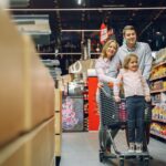 A parent with two young children navigating a grocery store aisle, looking at fresh produce, with a shopping cart full of healthy items.