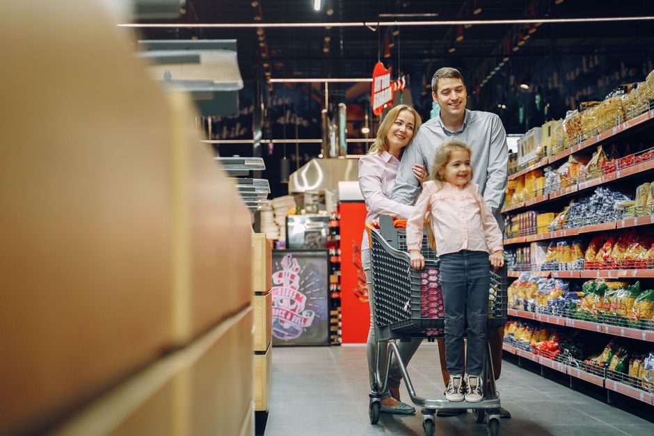 A parent with two young children navigating a grocery store aisle, looking at fresh produce, with a shopping cart full of healthy items.