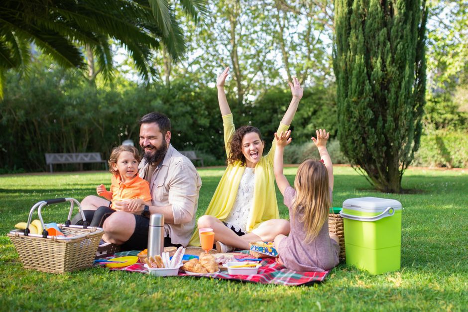 A family enjoying a picnic lunch on a sunny day in a large, green European city park, with historic buildings in the background.
