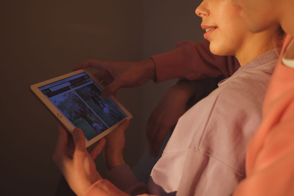 A family on a couch, mother, father, and child, happily viewing shared photos and videos on a large tablet, with other devices like a laptop and smartphone nearby, all connected digitally to a cloud icon.