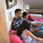 A family, including a parent and two children, sitting on a sofa and sharing a tablet, each looking at a different section of the screen.