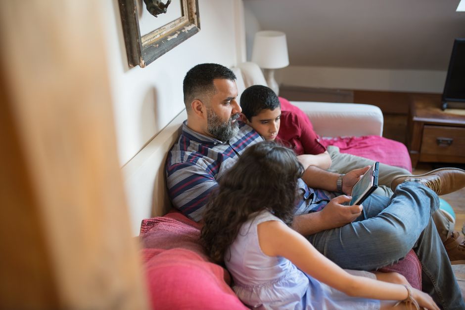 A family, including a parent and two children, sitting on a sofa and sharing a tablet, each looking at a different section of the screen.