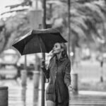 Stylish woman in a trench coat with an umbrella walking past a Parisian cafe in light rain.