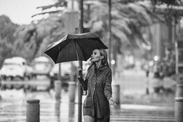 Stylish woman in a trench coat with an umbrella walking past a Parisian cafe in light rain.