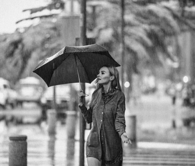Stylish woman in a trench coat with an umbrella walking past a Parisian cafe in light rain.
