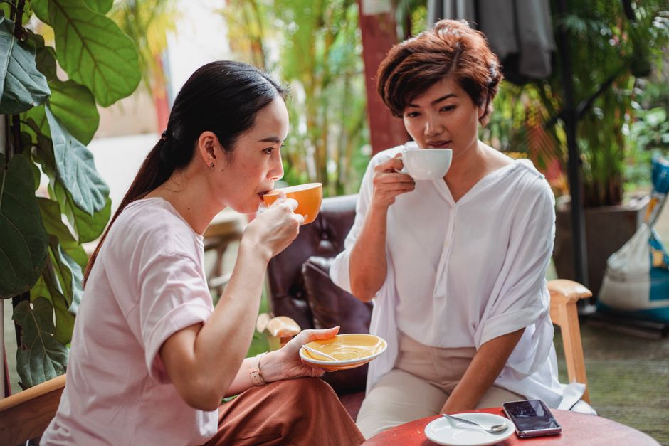 Two young people casually dressed, enjoying coffee and conversation at an outdoor cafe with historic architecture in the background.