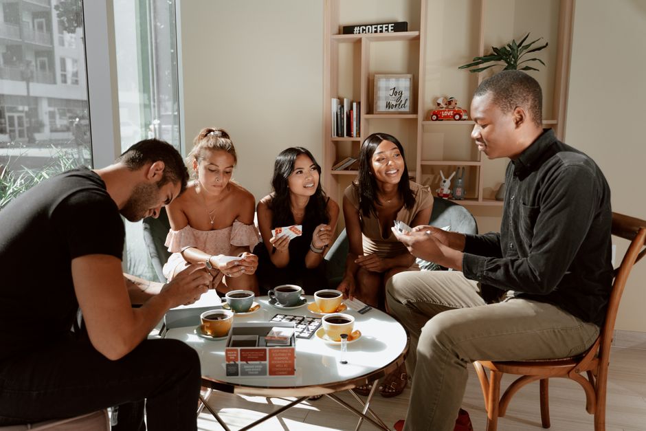 A group of friends laughing and interacting while playing a strategy board game, with game pieces and cards spread across the table.