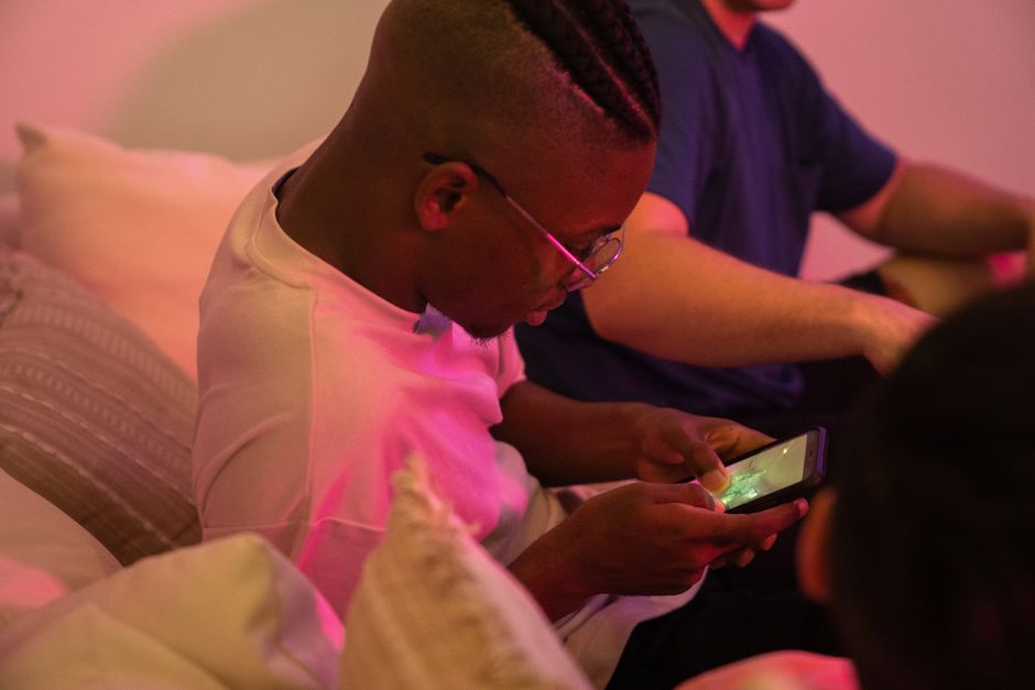 Diverse group of young adults and teens gathered around, intensely focused on their mobile devices, playing a multiplayer creative building game together.