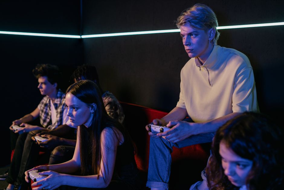 Three friends gathered in a living room, each focused on their respective gaming platforms: one on an Xbox, another on a PlayStation, and a third on a Nintendo Switch, depicting diverse gaming habits and preferences.