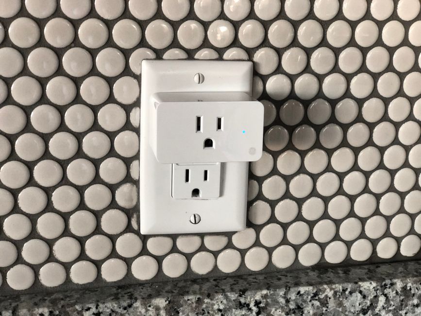 Close-up of a hand plugging a lamp into a smart plug connected to a wall outlet in an apartment.