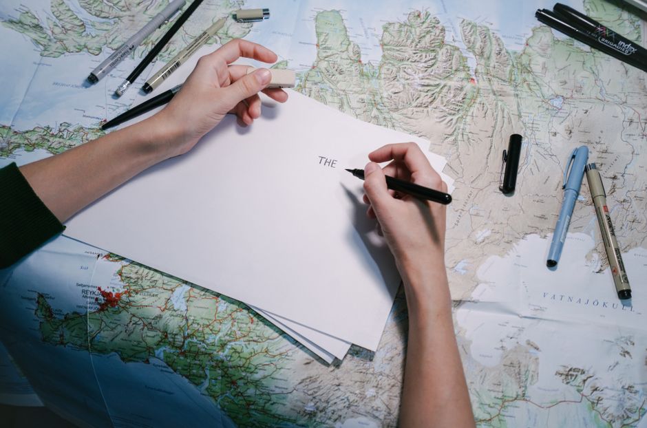 Two children, perhaps siblings, sitting at a table with a large map spread out, pointing and drawing on it with colorful markers, actively participating in trip planning.