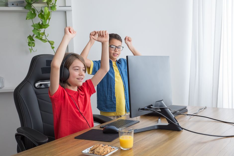 Two children are seen laughing and interacting while playing Roblox on separate computers, with chat bubbles above their screens, representing online communication and interaction in games.