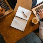 A person working on a silver laptop at a café table with a cup of coffee and notebook