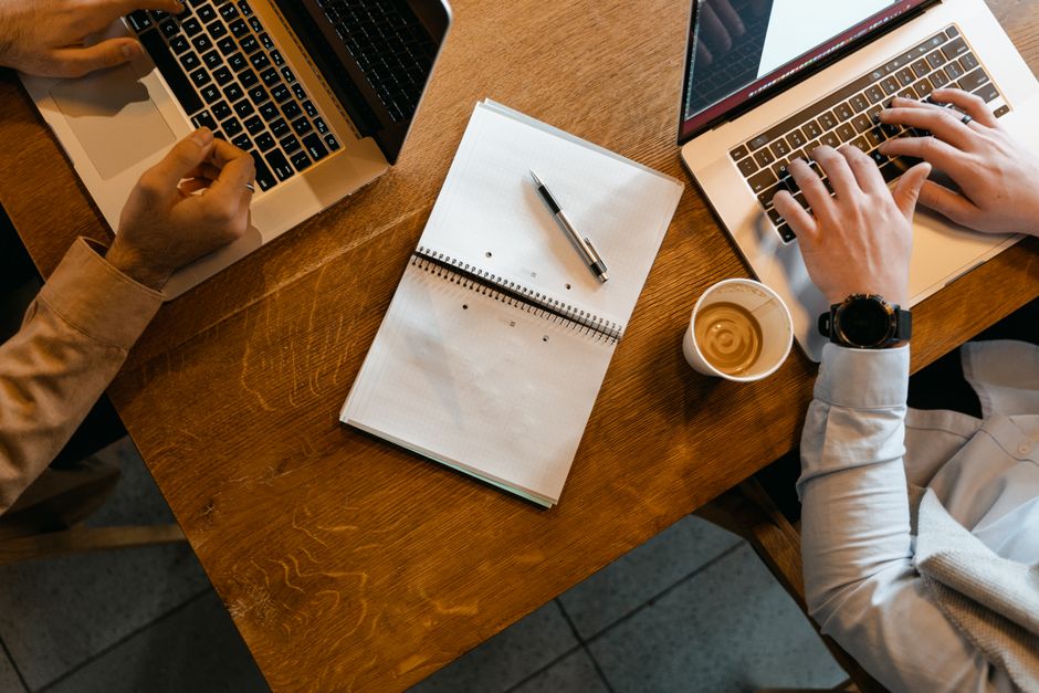 A person working on a silver laptop at a café table with a cup of coffee and notebook