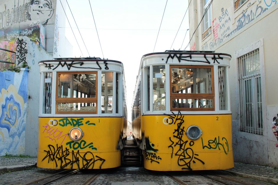 A historic yellow tram in Lisbon winding through a hilly, colorful neighborhood, with a glimpse of the Tagus River in the background.