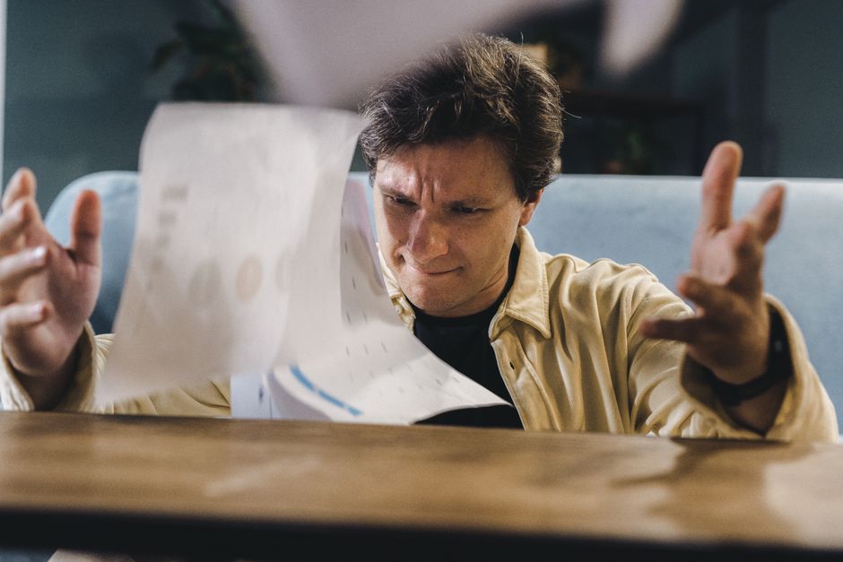 A person with a frustrated expression sitting at a chaotic desk overflowing with scattered papers and documents.
